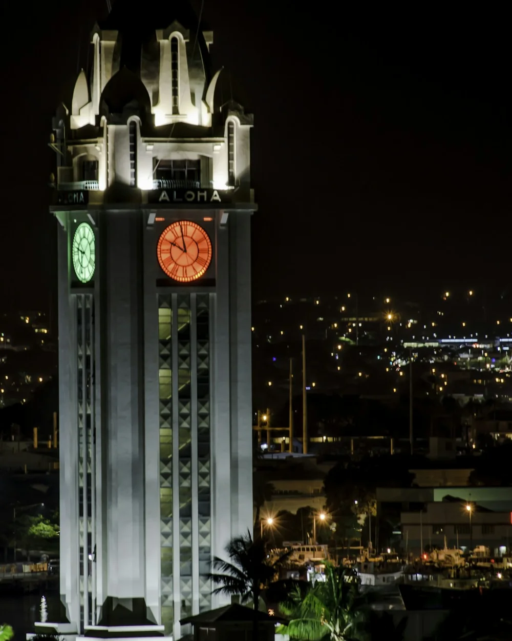 Historická hodinová věž Aloha Tower osvětlená v noci nad přístavem Honolulu, její ciferníky a nápis „ALOHA" zářící proti tmavé obloze