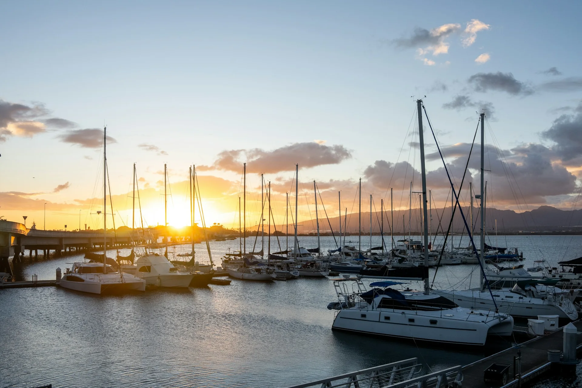 Havajské marina při západu slunce, stěžně plachetnic silhouetované proti zlaté a růžové obloze nad vodou (reprezentativní fotografie přístavu v oblasti Honolulu při západu slunce, nikoli vlastní trávník Ka Moana)